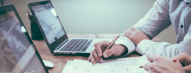 Zwei angeschnittene Personen sitzen am Schreibtisch bei geöffneten Laptops